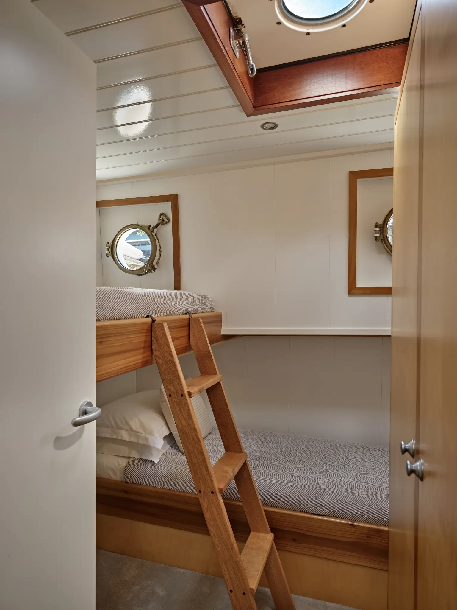 Cozy bedroom corner with a green pillow, white bedding, wooden nightstand with flowers, lit wall lamp, and a window showing waterfront buildings and boats.