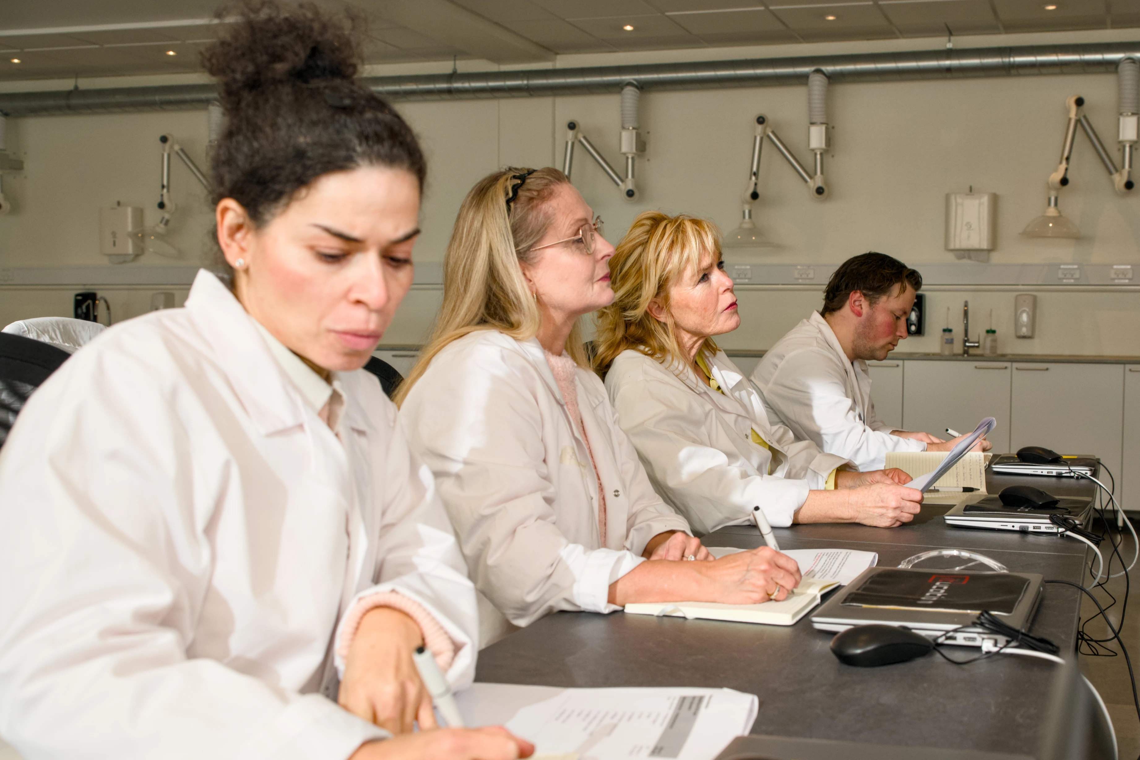 Fire kursister i hvide kitler tager noter under GMP undervisning i et laboratorielokale
