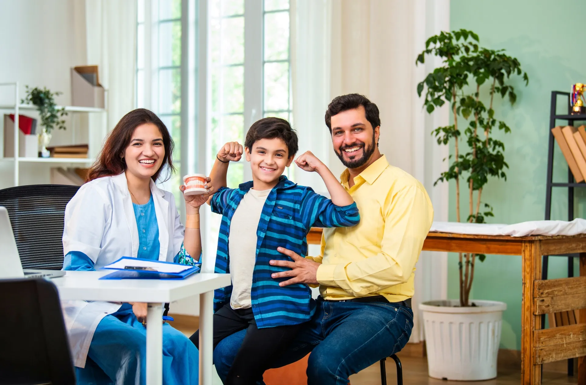 Smiling dentist, boy flexing muscles, and man sitting together in a bright dental office.