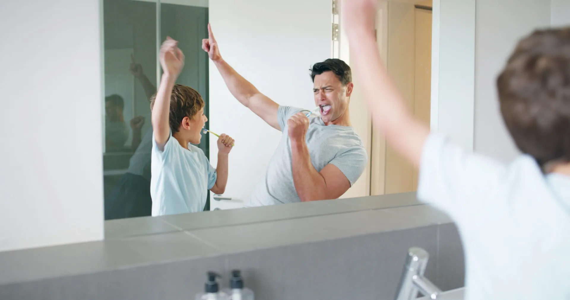Father and son brushing teeth together in bathroom mirror, happily raising hands and singing.