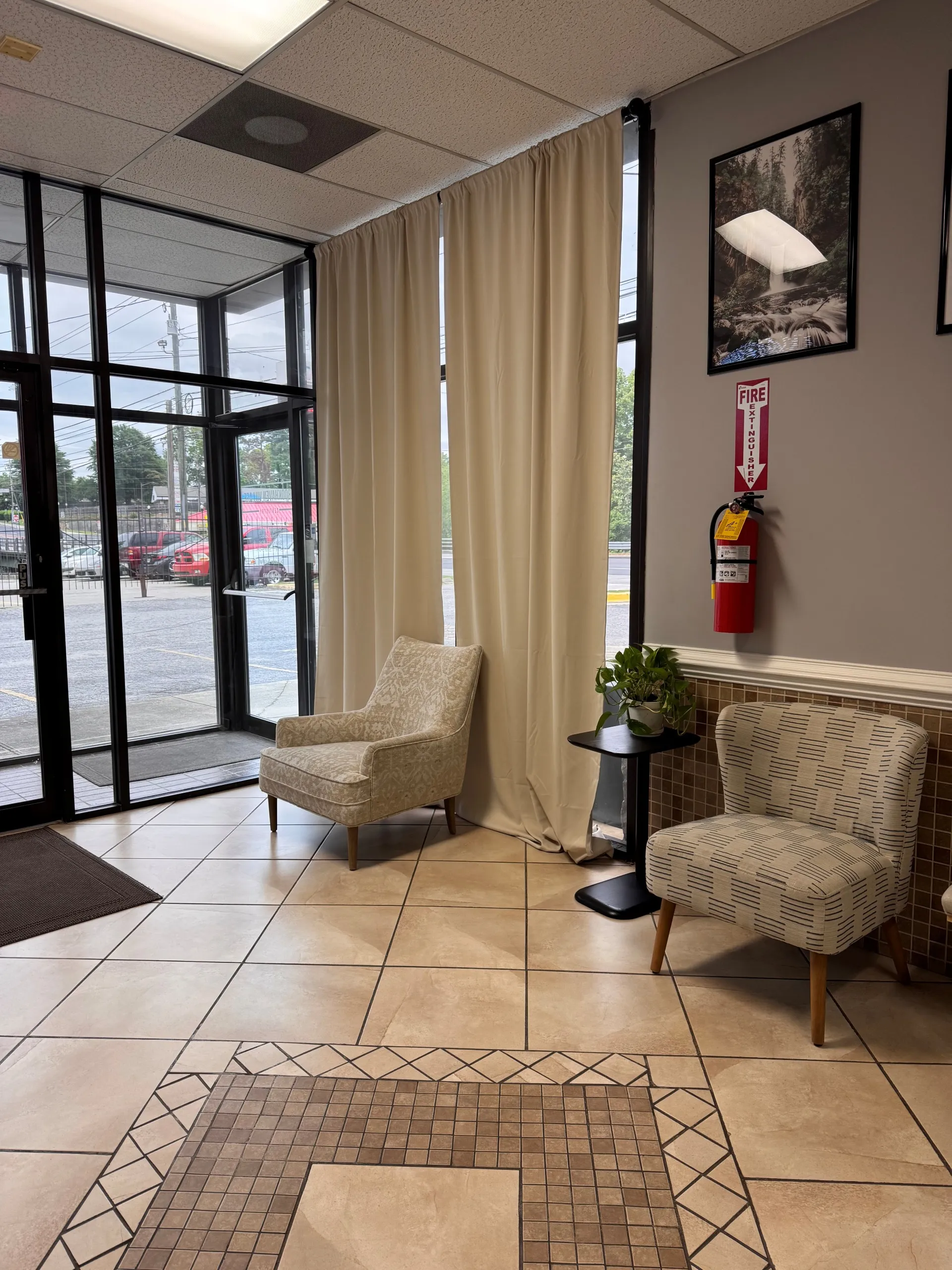 Waiting area with two patterned chairs, a small table with a plant, beige curtains, and a fire extinguisher mounted.
