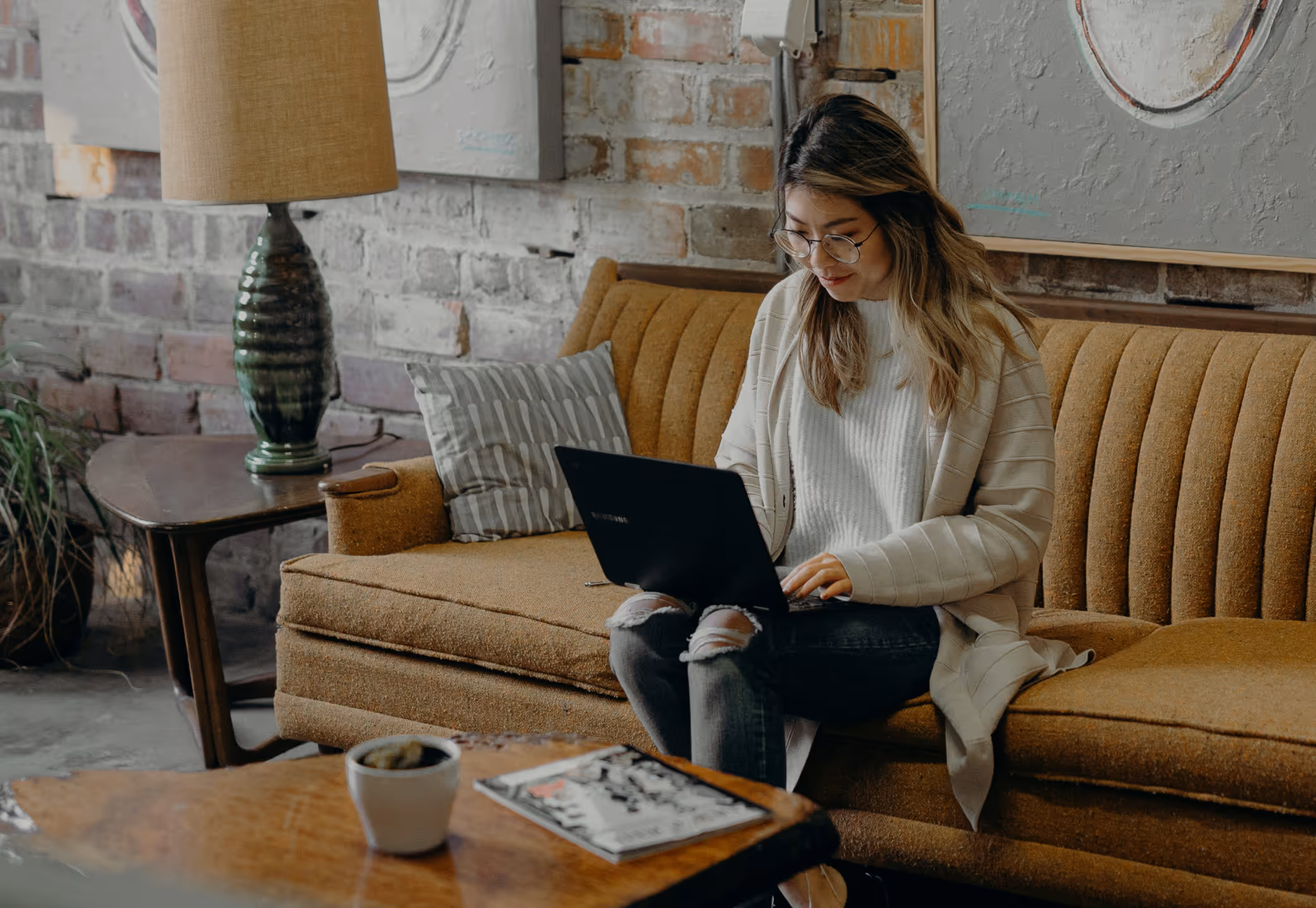 A woman sits on a beige couch working on a laptop
