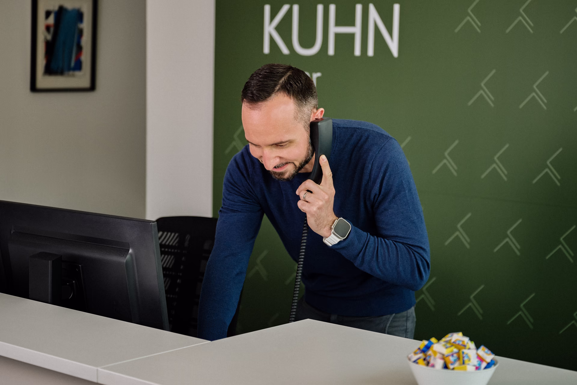 Mann am Empfang telefoniert, lehnt sich vor PC, grüner Hintergrund mit Schrift KUHN.