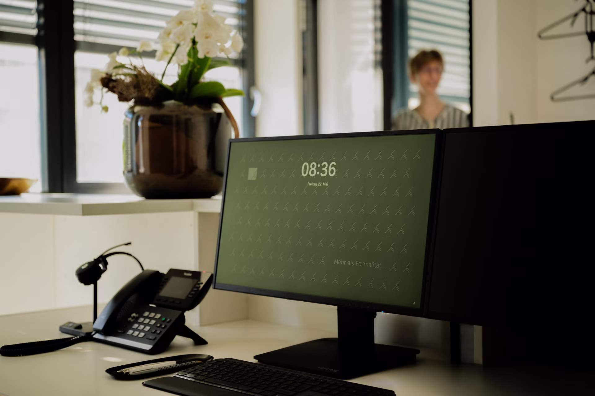 Bürotisch mit Computerbildschirm, Telefon und einer Vase mit weißen Blumen im Hintergrund.