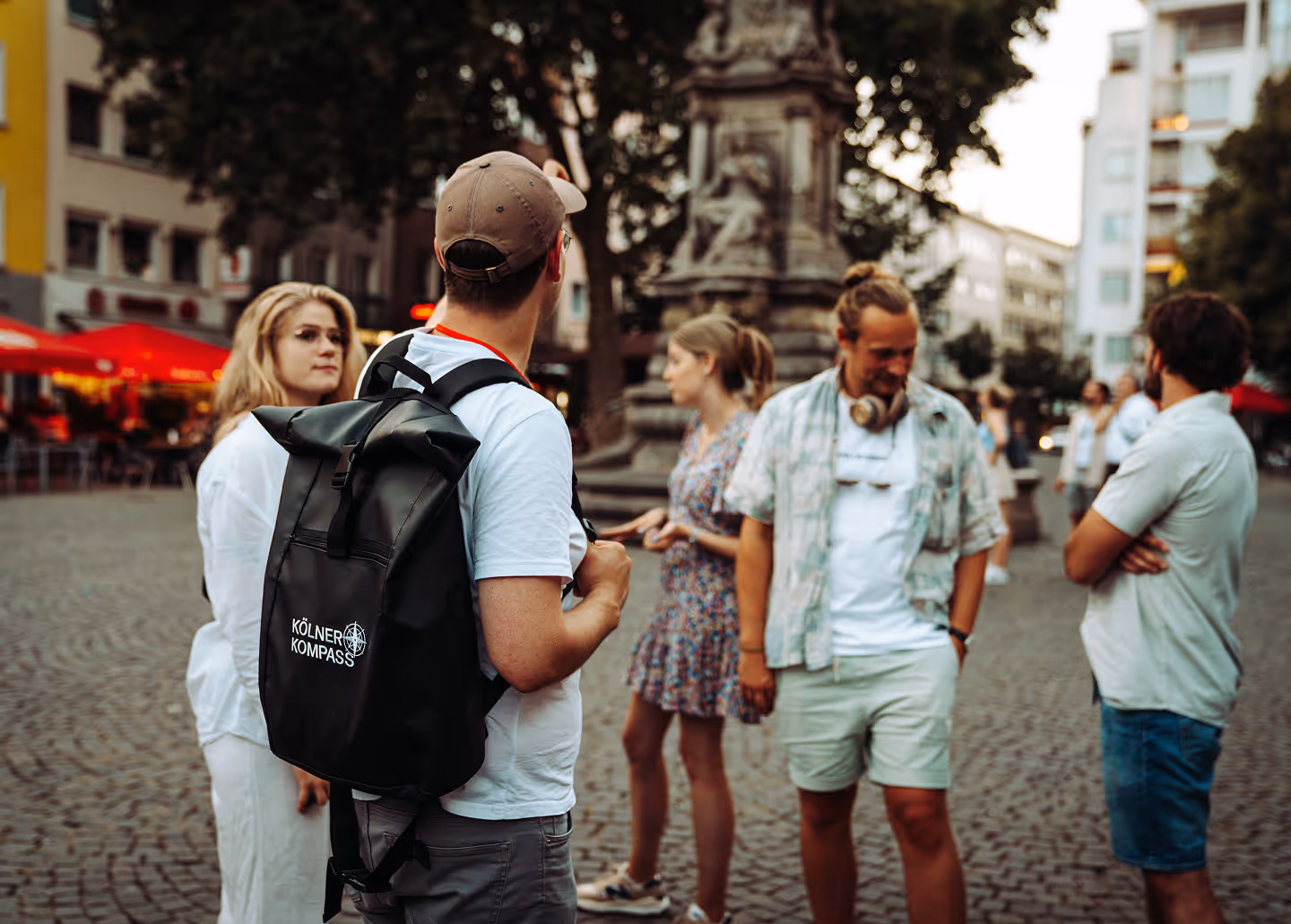 Kölner Kompass Tourguide von hinten mit Rucksack erzählt Gästegruppe auf einem Kölner Platz