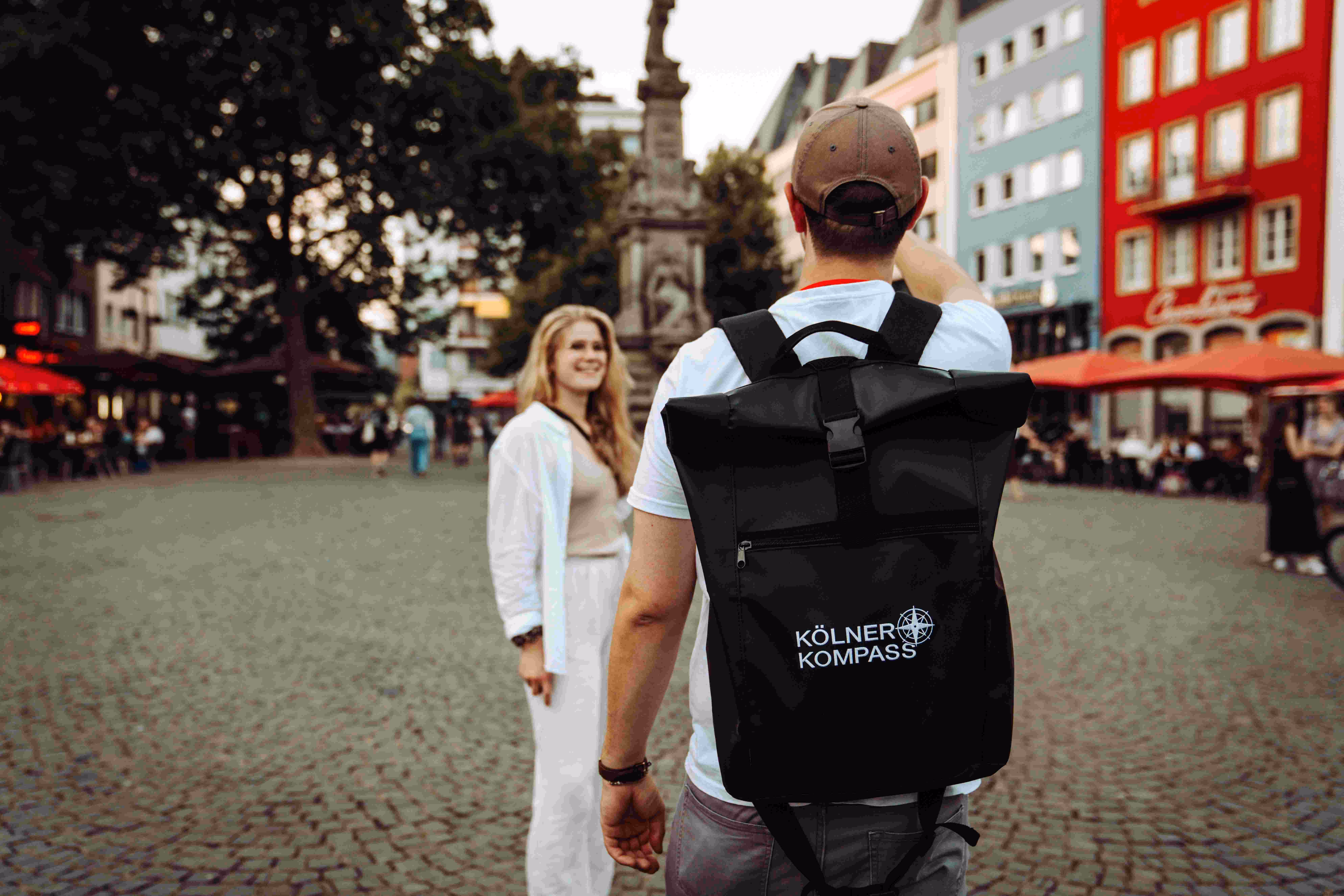 Kölner Kompass Tourguide von hinten mit Rucksack führt Gast über einen Platz in der Kölner Altstadt