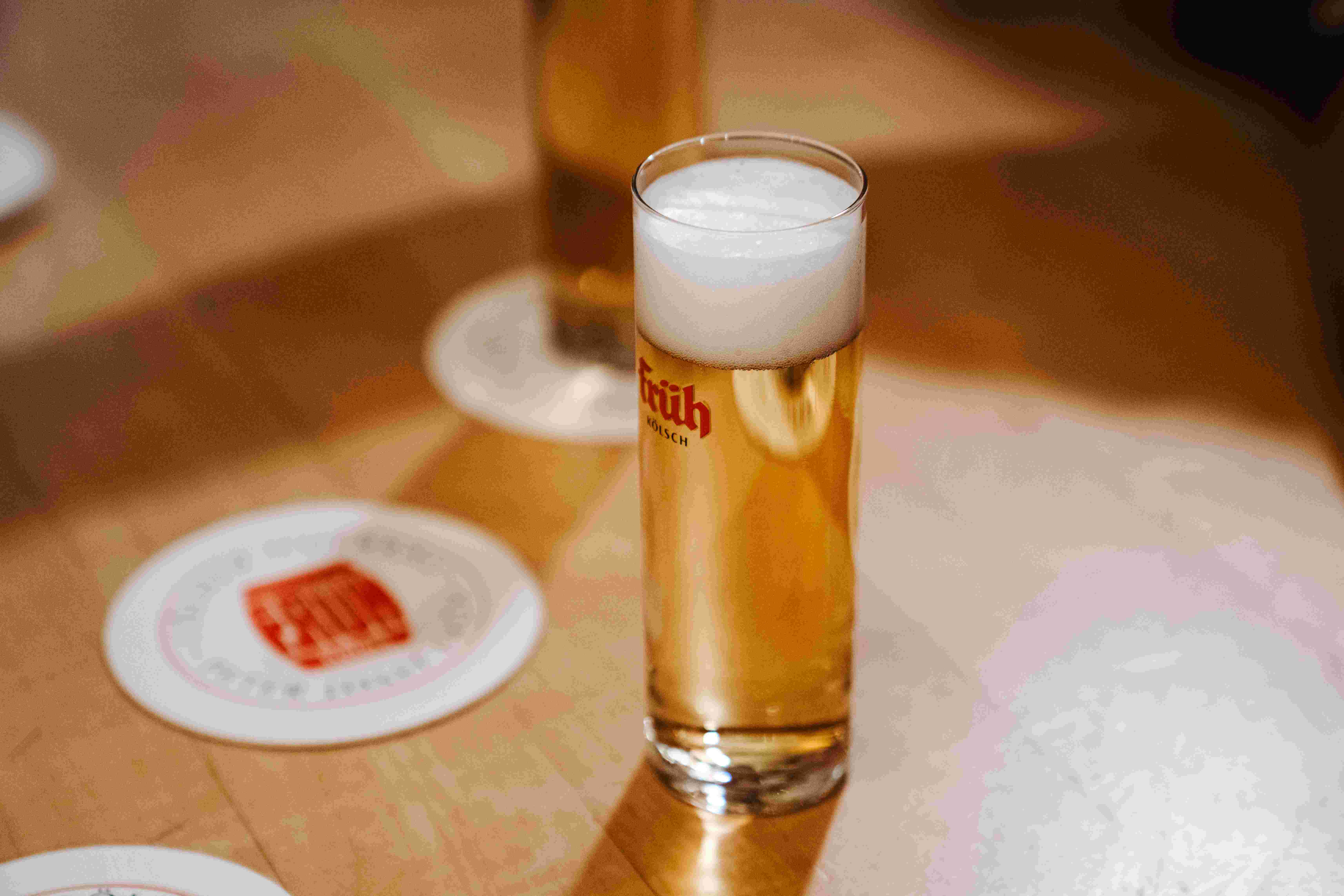 Ein Glas Früh Kölsch Bier mit Schaum steht auf einem Holztisch neben Bierdeckeln.