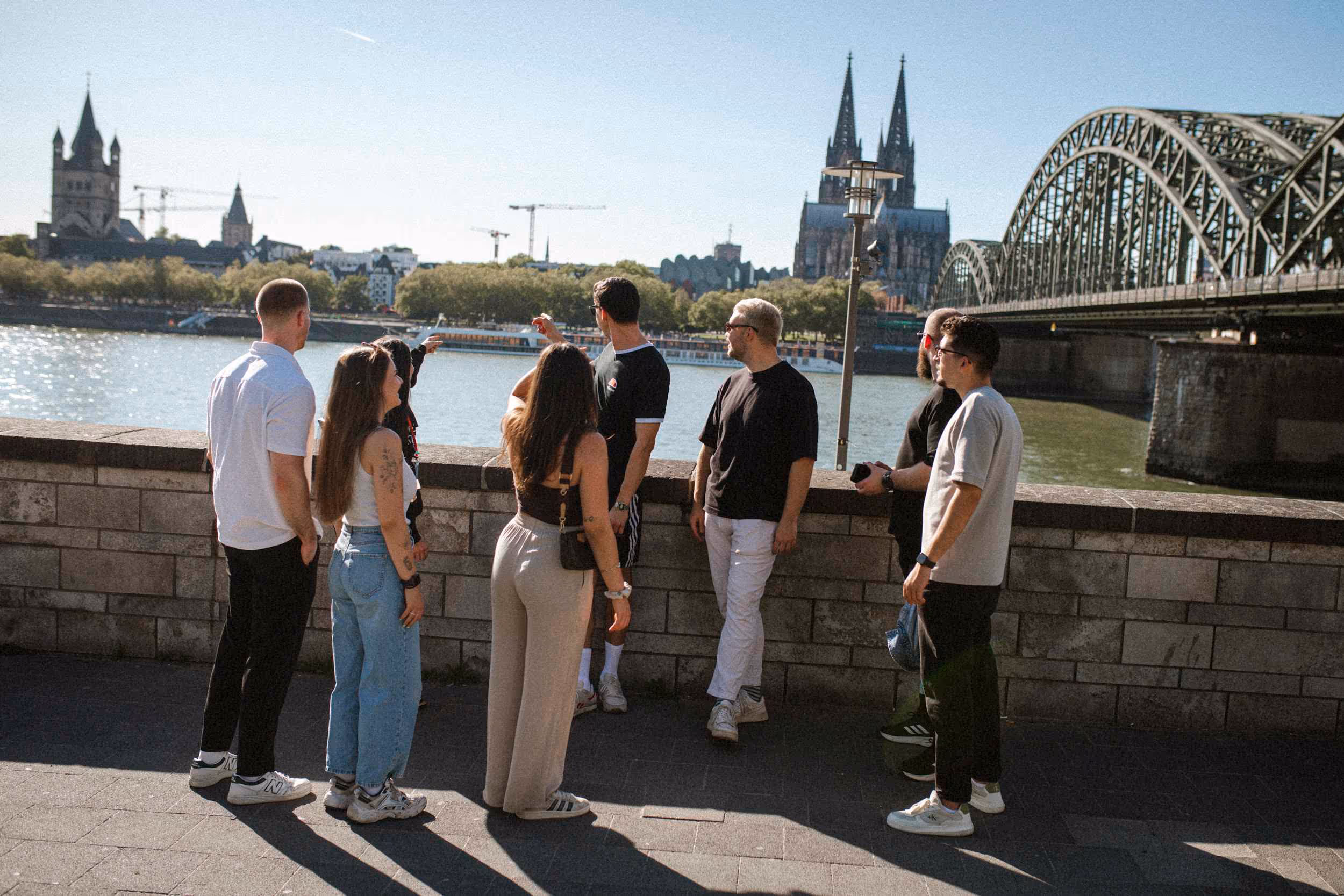 Kölner Kompass Tourguide steht mit Gästegruppe am Rheinufer mit Blick auf Dom und Hohenzollernbrücke