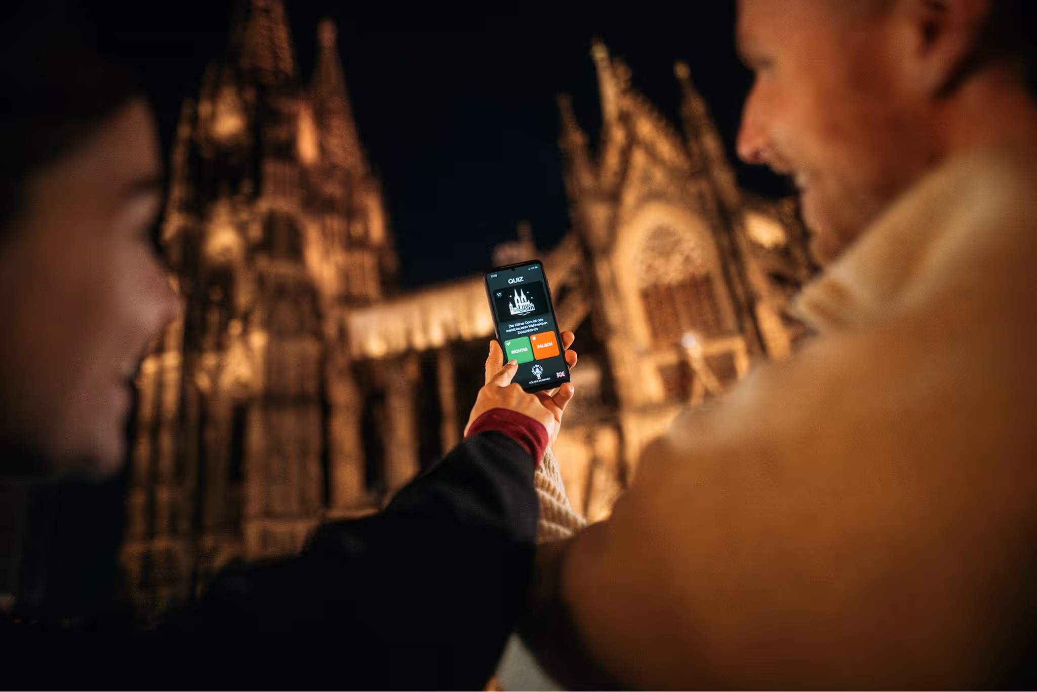 Zwei Personen schauen sich die Kölner Kompass App vor dem beleuchteten Dom bei Nacht an