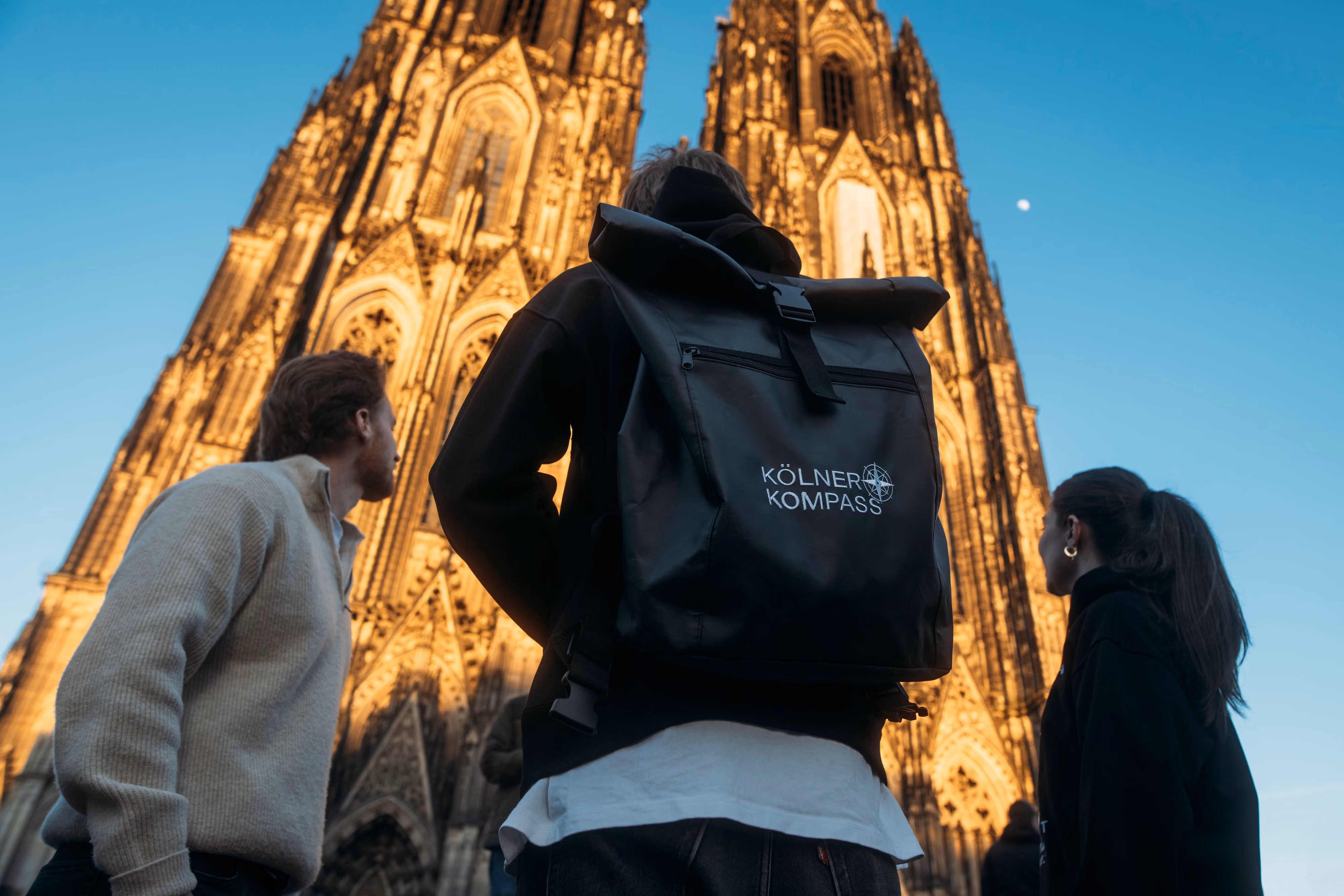 Kölner Kompass Tourguide mit Rucksack steht vor dem beleuchteten Kölner Dom in der Abenddämmerung