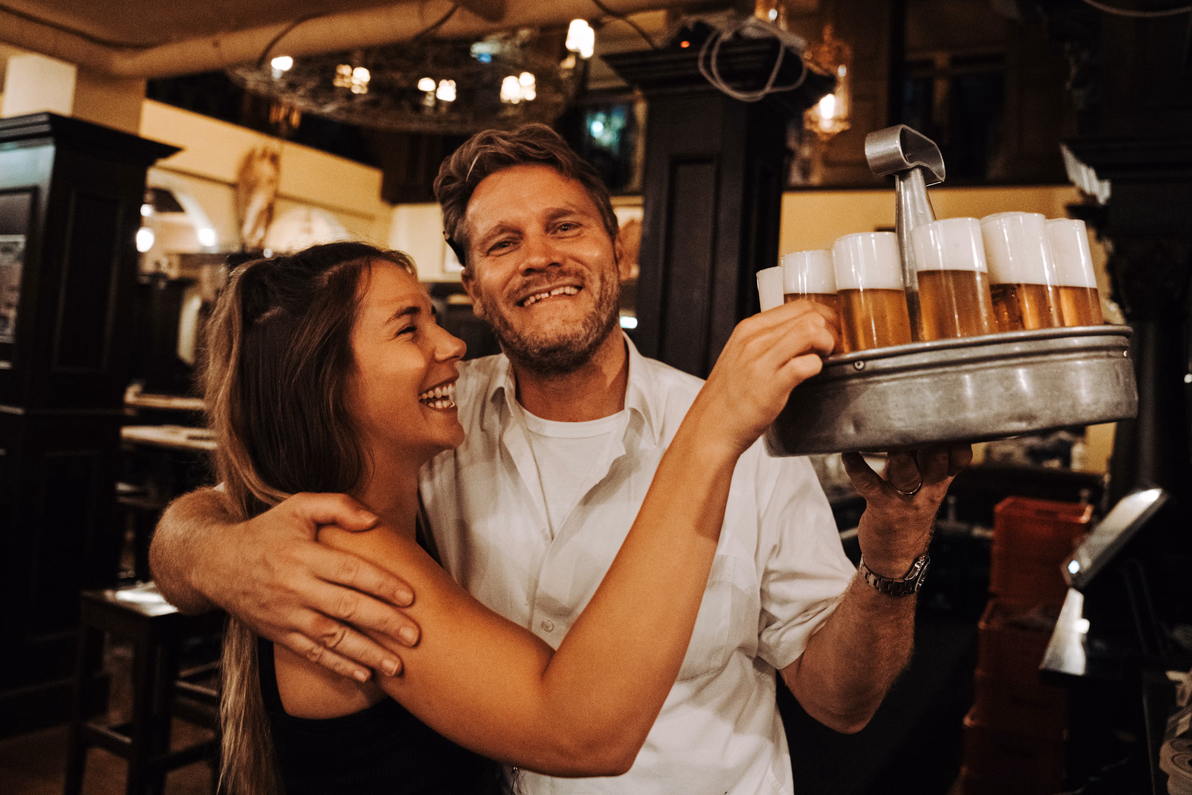 aar umarmt sich lachend, während der Mann ein Tablett mit Kölsch-Gläsern in einem Kölner Brauhaus hochhält