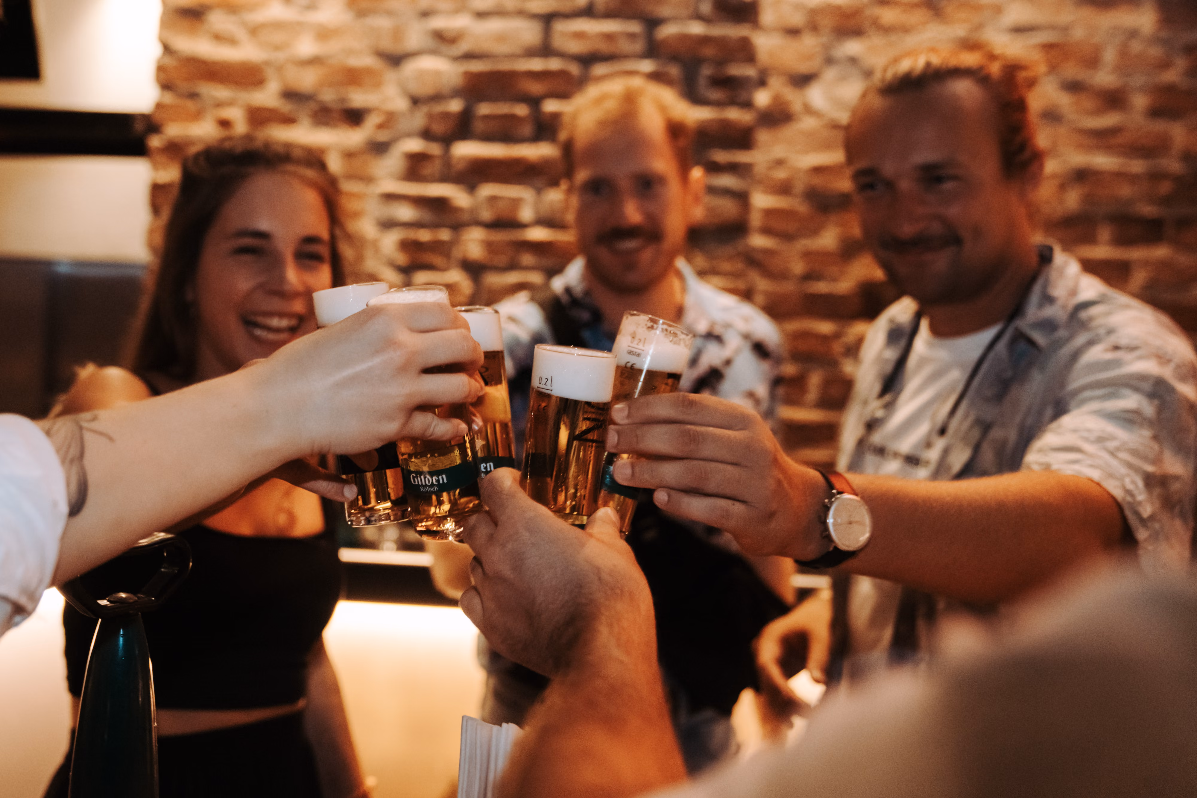 Gruppe stößt lachend mit Kölsch-Gläsern vor einer Backsteinwand in einem Kölner Brauhauskeller an