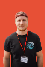 Junger Mann mit rotem Cap, schwarzem T-Shirt und Namensschild vor rotem Hintergrund.