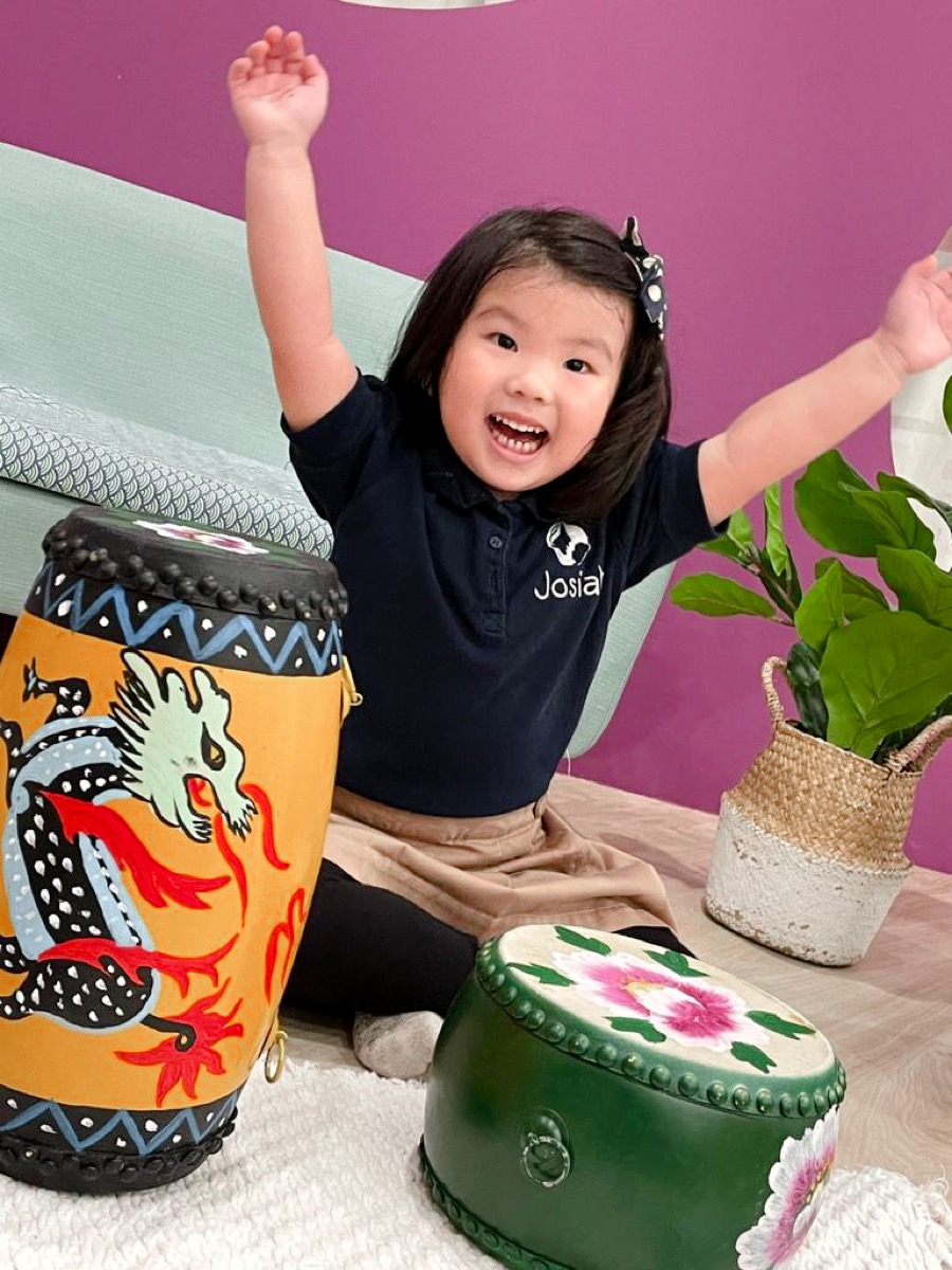 A delighted child throwing her arms up with joy beside hand-painted drums at Josiah Music Lab