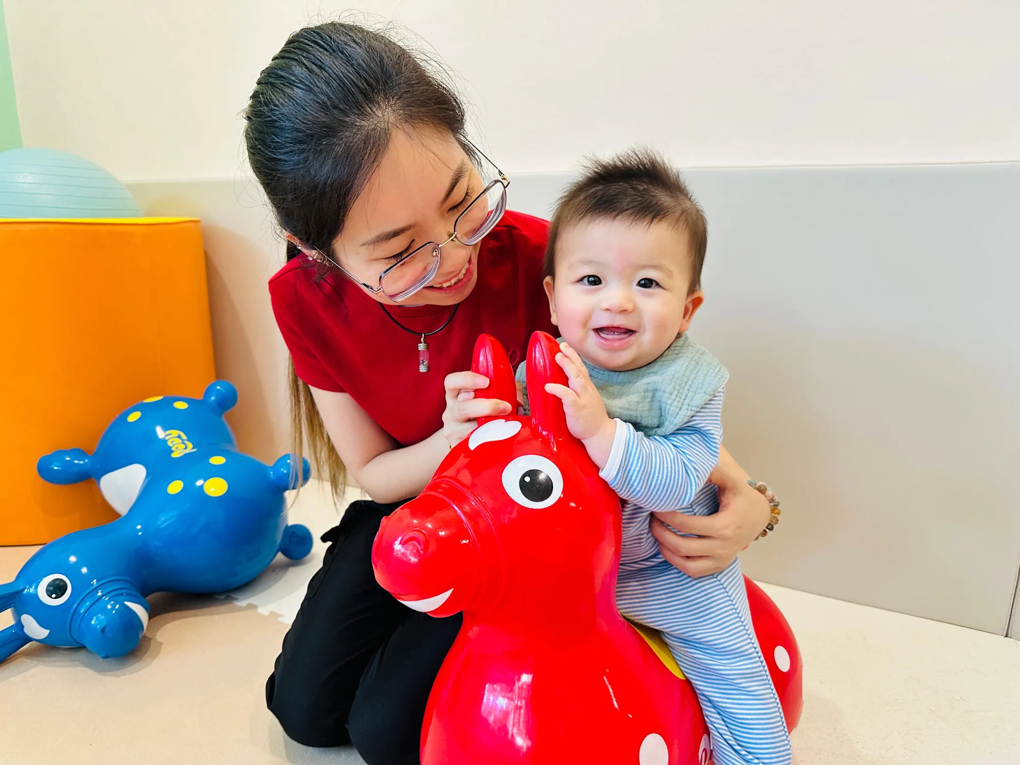 A Josiah Montessori educator kneeling on the floor beside a baby on a ride-on toy