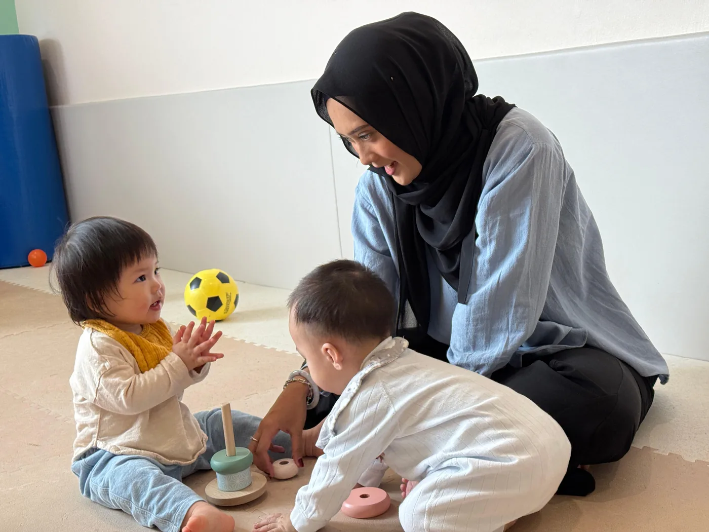 A Josiah educator on the mat with two babies and a wooden Montessori stacking toy