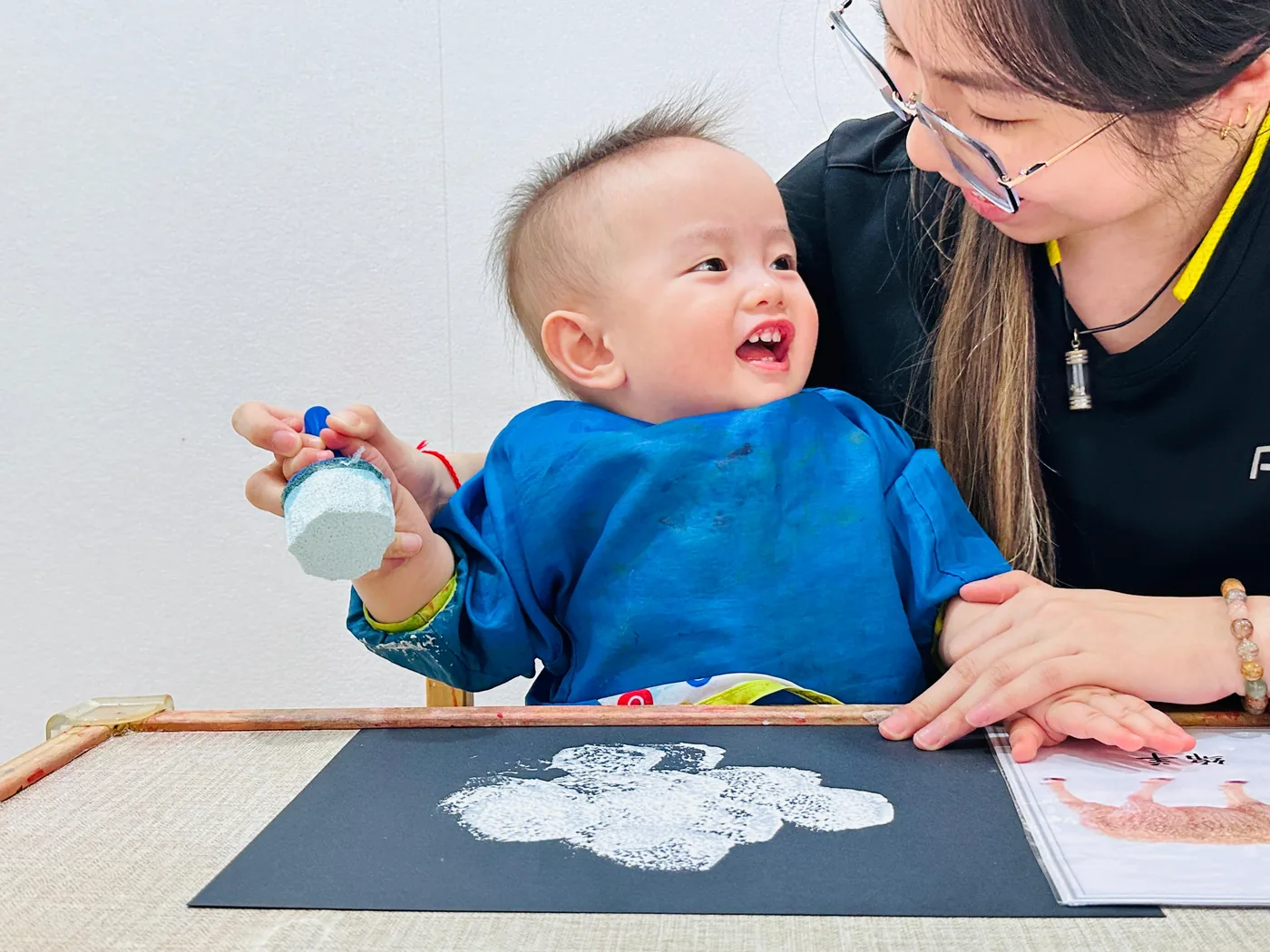 A baby printing with a sponge on black paper while a Josiah educator sits beside him