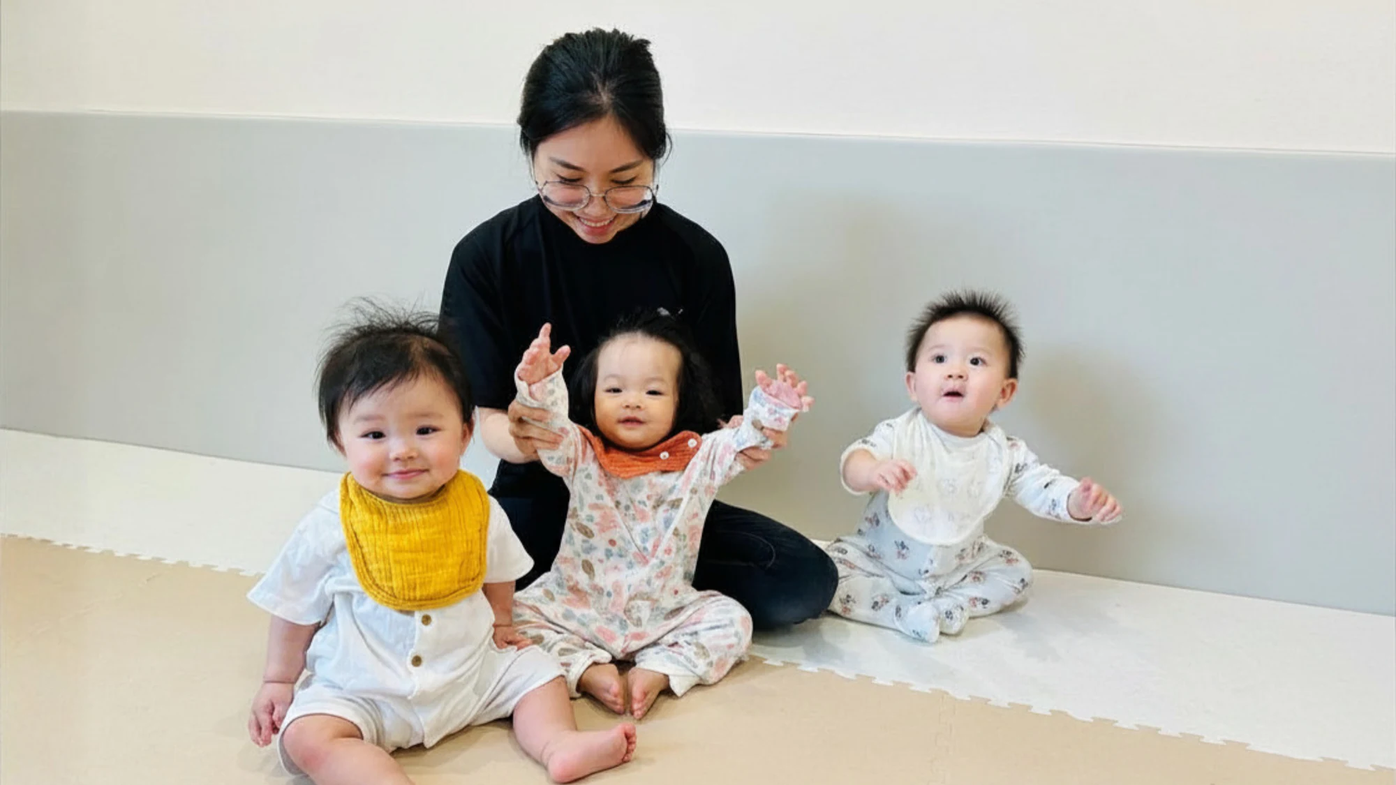 Three babies sitting in a row on the mat in a Josiah Montessori infant room, an educator kneeling behind them