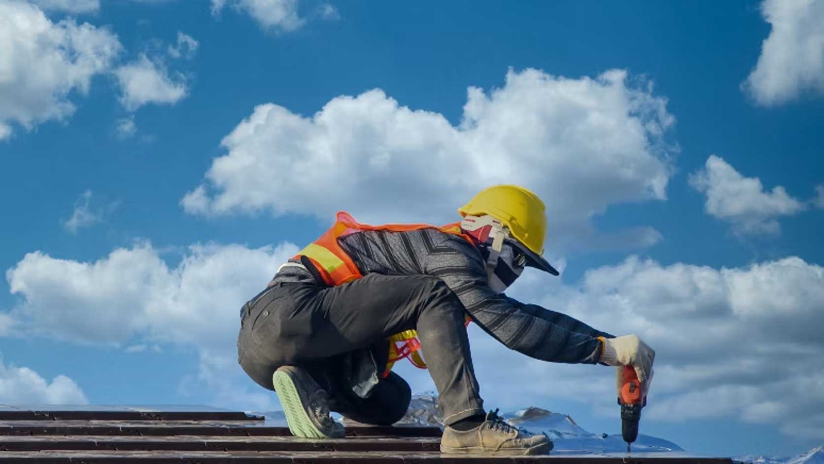 Professional roofer wearing safety equipment using a power drill while carrying out roofing work under a clear blue sky.