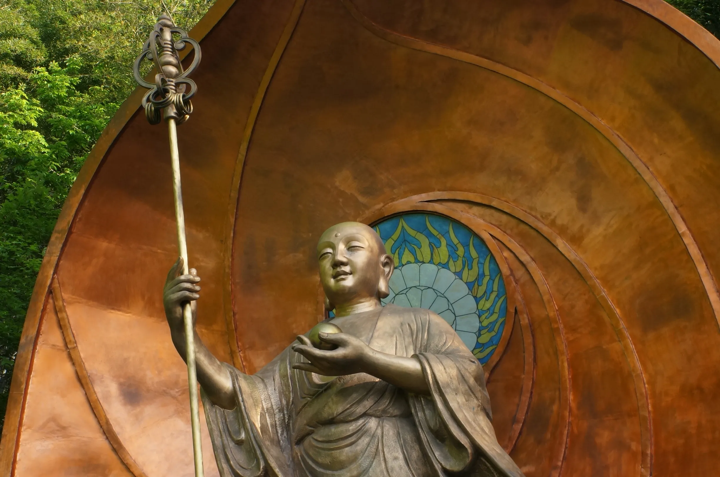 Patinated Buddhist figure holding a staff before a circular bronze backdrop.
