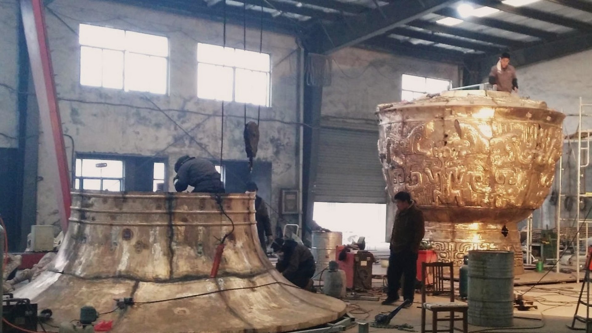 Workers position large decorated metal vessel sections for assembly in the workshop.