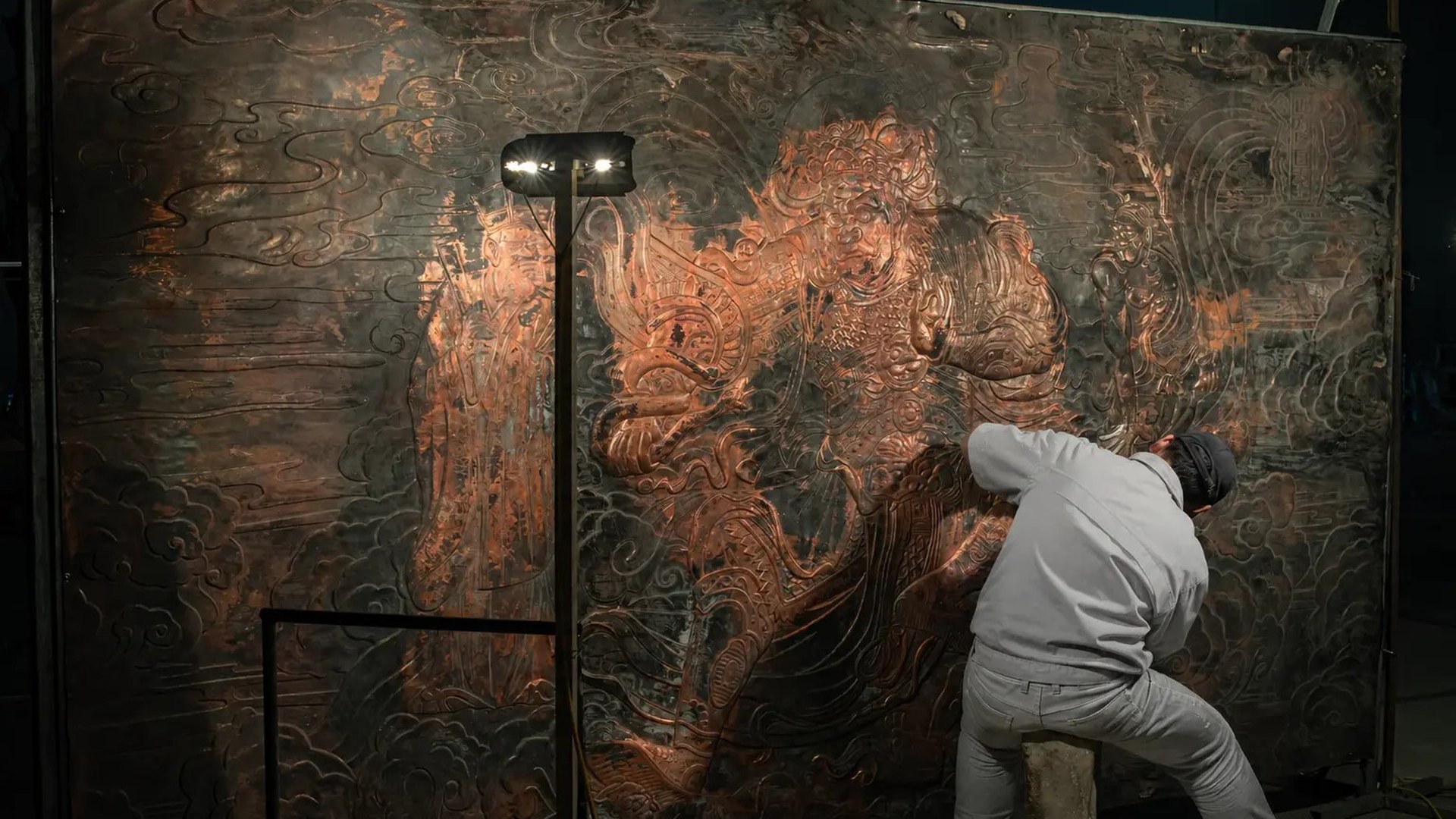 Artisan hand-chases detail across a monumental copper relief panel.