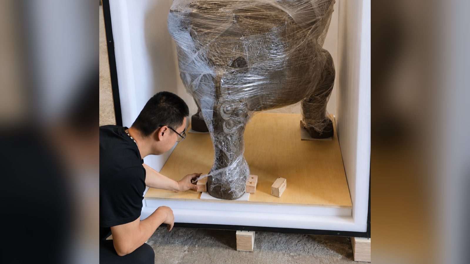 A worker fitting a timber support beside the foot of a wrapped vessel inside a crate.