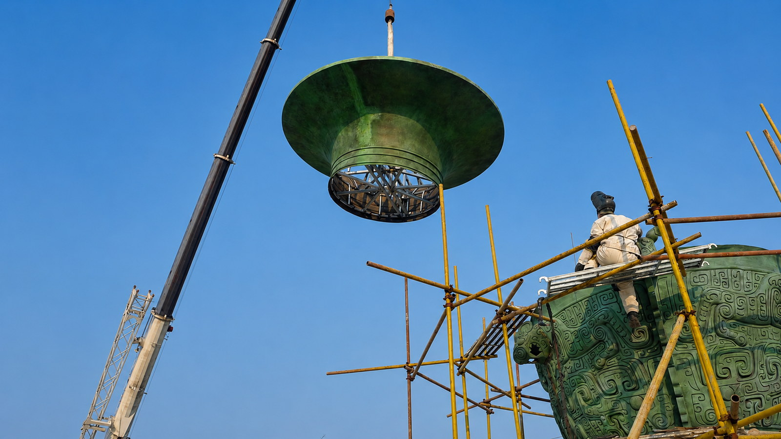 A crane lifting the flared upper section of a large bronze vessel above a work platform.
