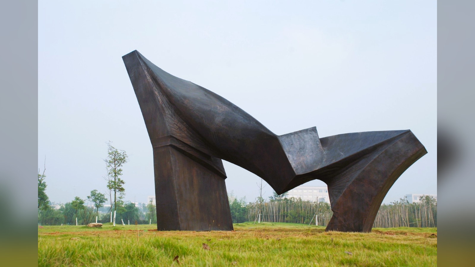 An abstract metal sculpture installed on a lawn with trees and buildings in the background.