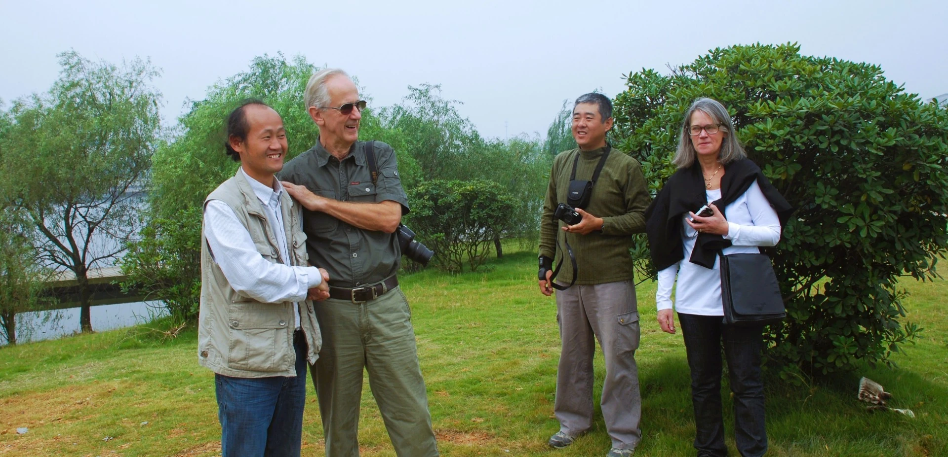 Four people discuss sculptures beside a landscaped lakeside area.