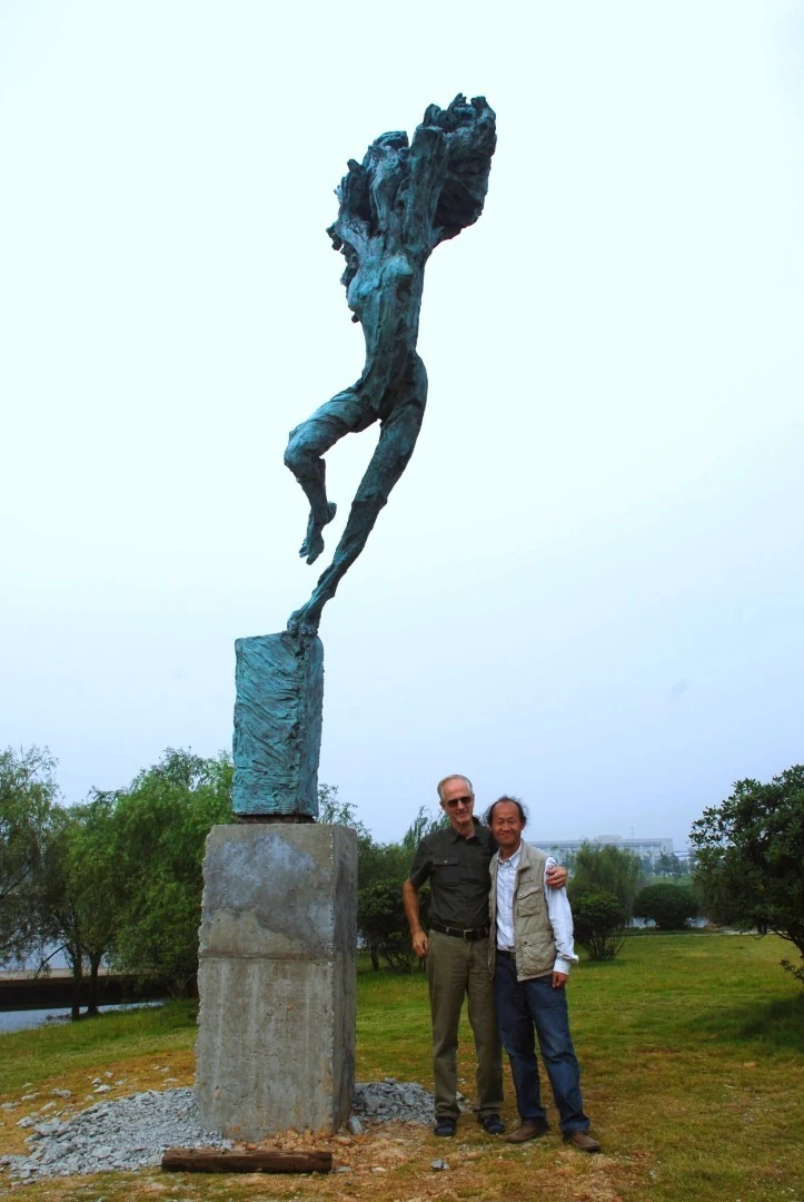 Two people stand beside a tall outdoor figurative sculpture.