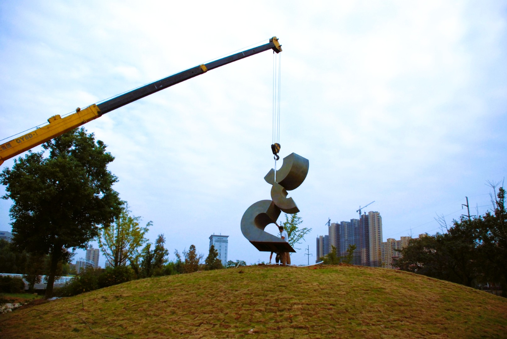 A crane positions an abstract sculpture on a grassy mound.