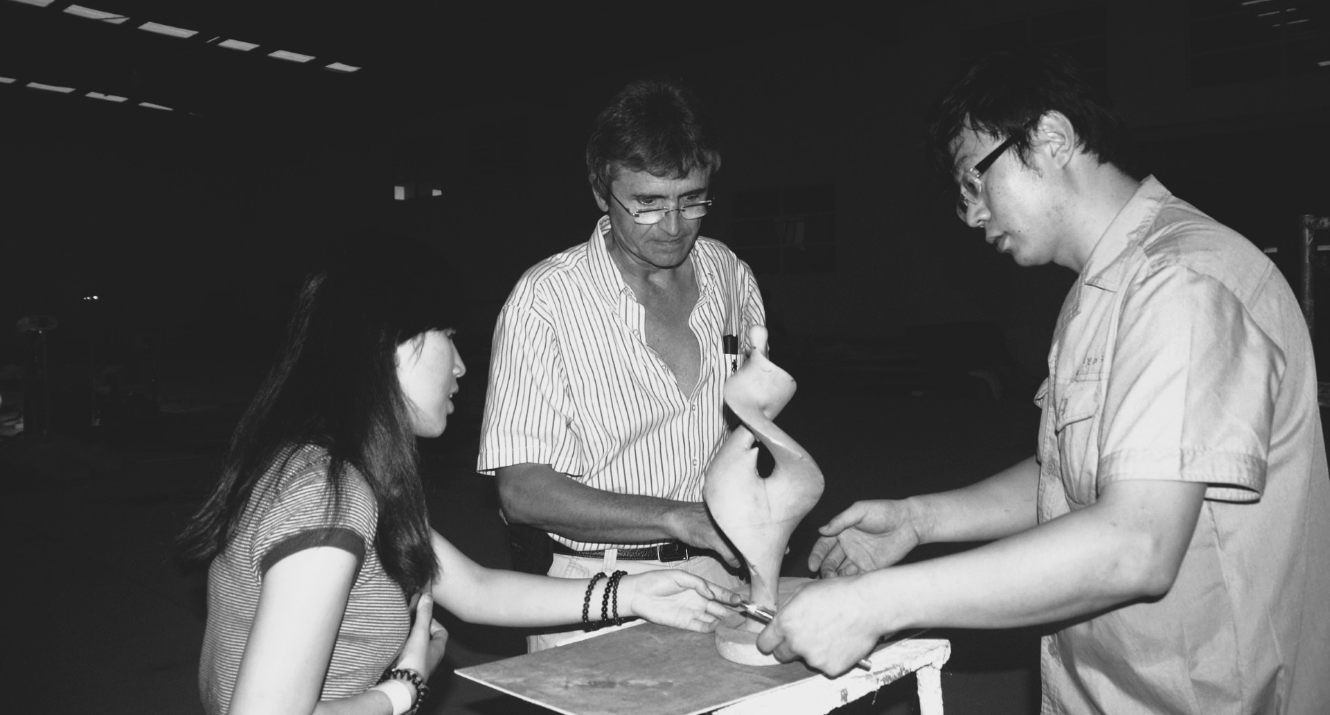 Black-and-white photograph of three people discussing a small sculpture model in the workshop.