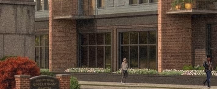 Two people walk on sidewalk outside brick building with flowers near the entrance.