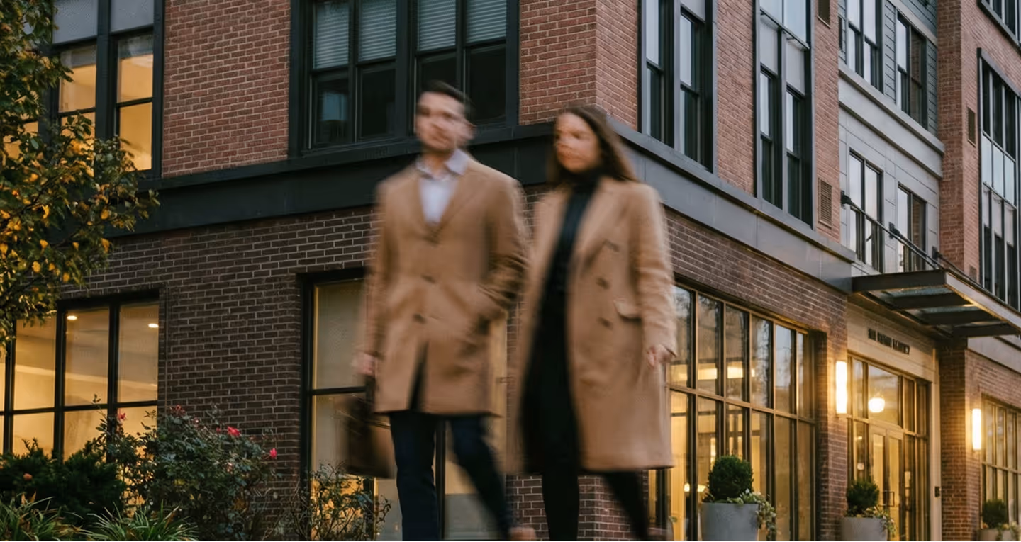 Blurred image of man and woman walking past a modern brick building with large windows and plants.