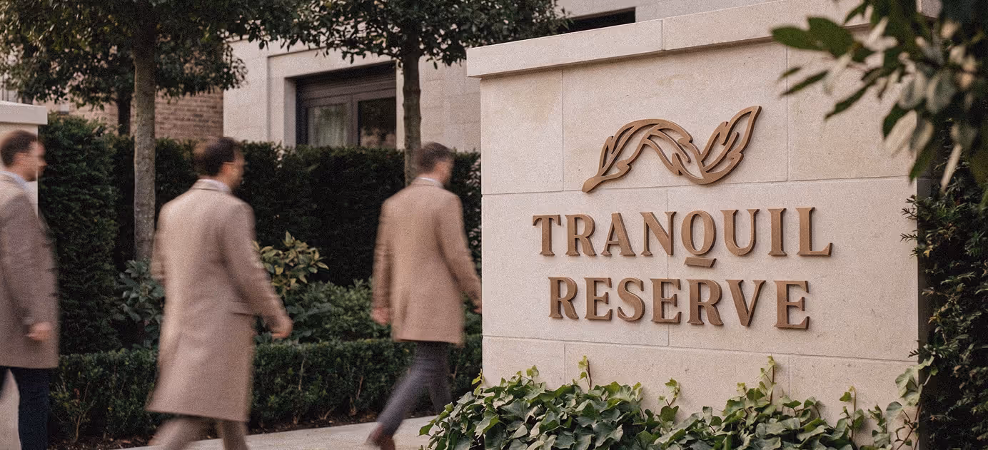 Three men in beige coats walking past a stone wall with Tranquil Reserve sign and leaf logo.