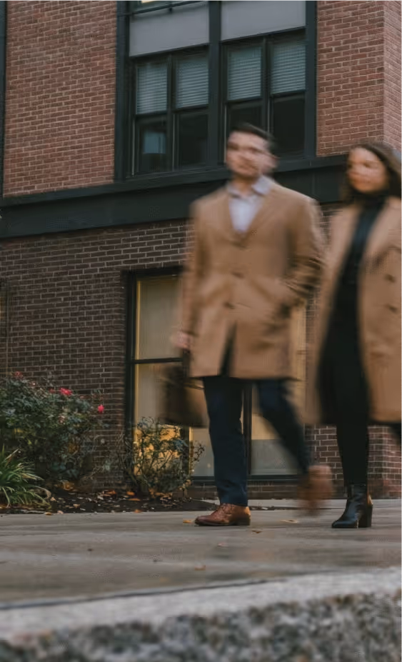 Blurred image of man and woman walking past a modern brick building with large windows and plants.