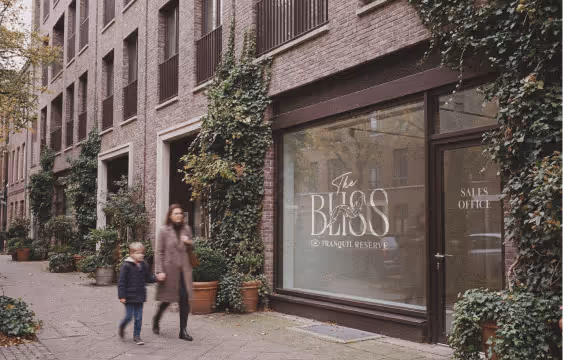 Woman and child walking past a brick building with a sales office window for The Bliss Tranquil Reserve.