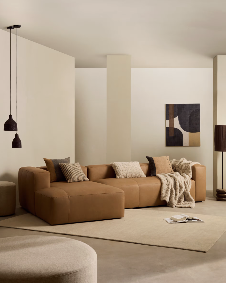 Modern living room with tan leather sectional sofa, pillows, fur throw, beige rug, and abstract wall art.
