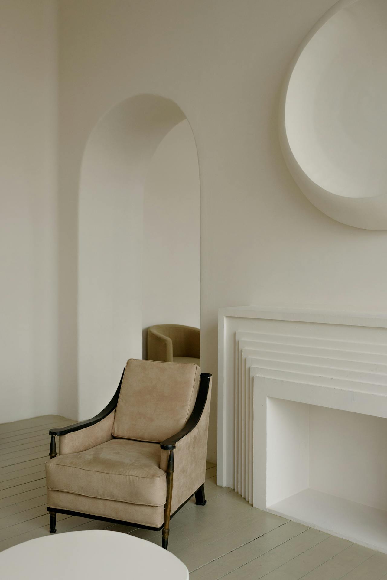 Beige armchair with black wooden arms in a minimal room with arched mirror and geometric fireplace.