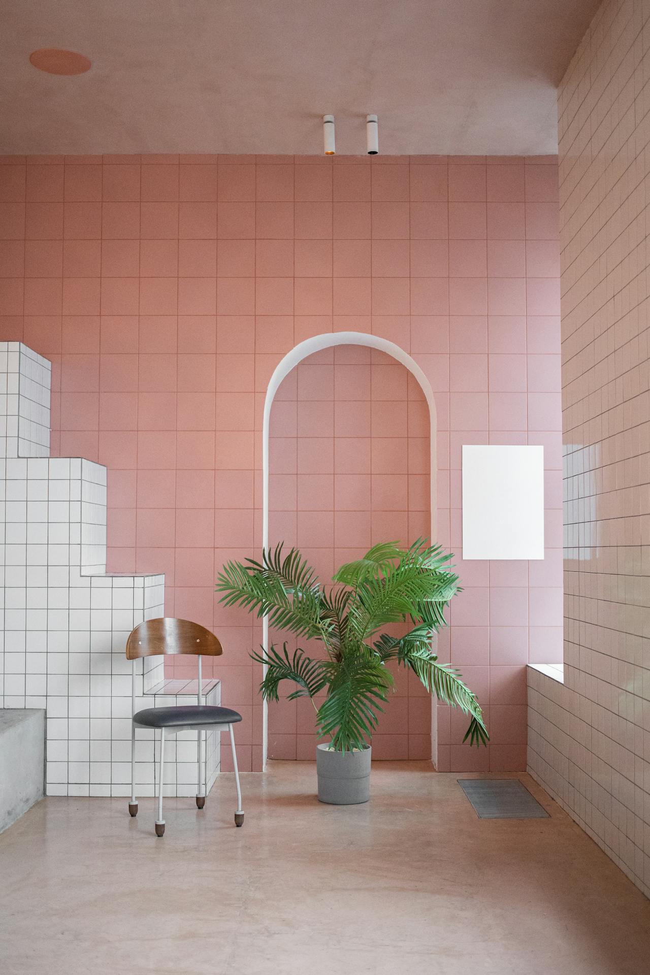 Minimalist pink tiled room with an arch, potted palm plant, vintage chair, and steps tiled in white.