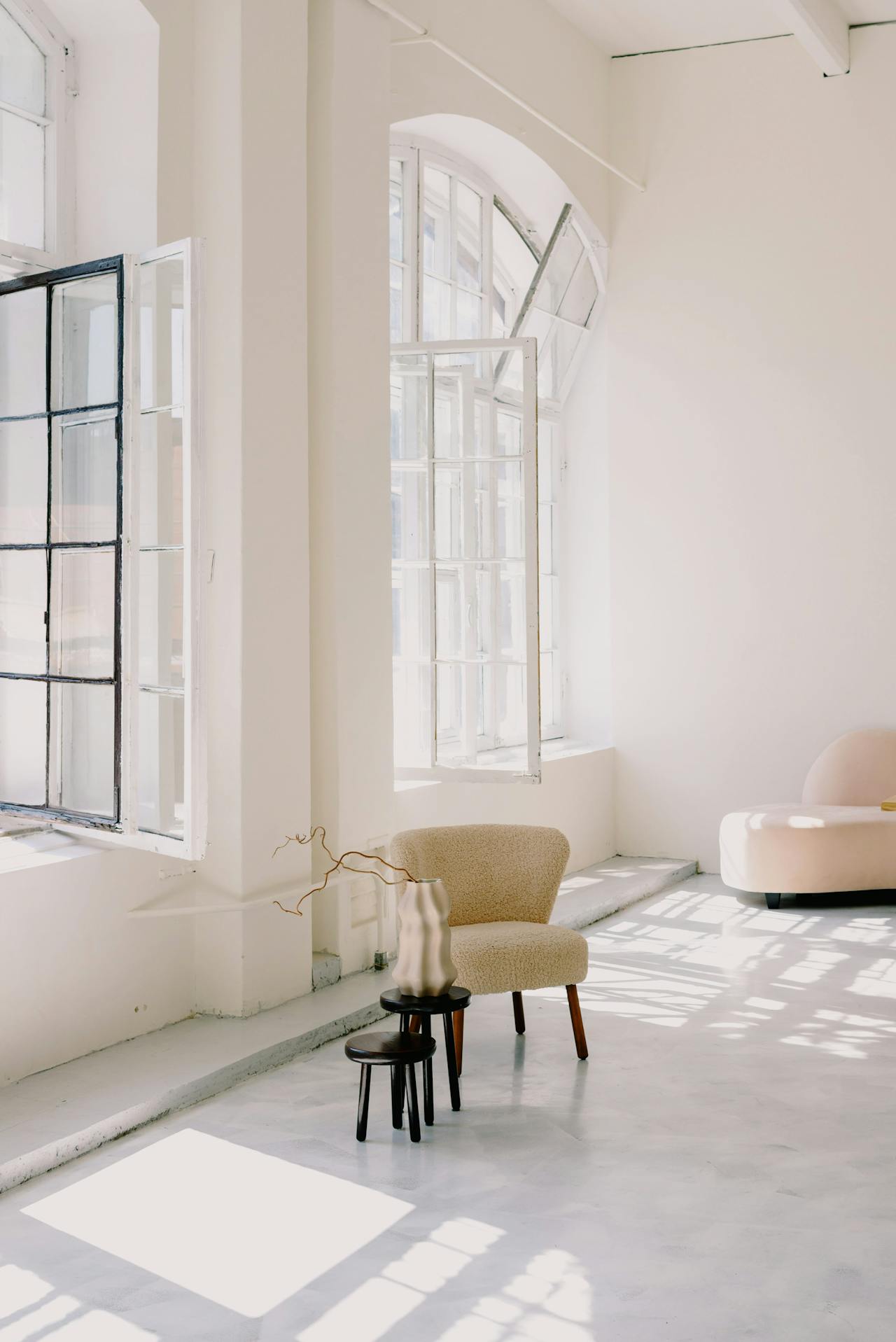 Bright minimalist room with open windows, a beige chair, two small tables, and a sculptural vase.