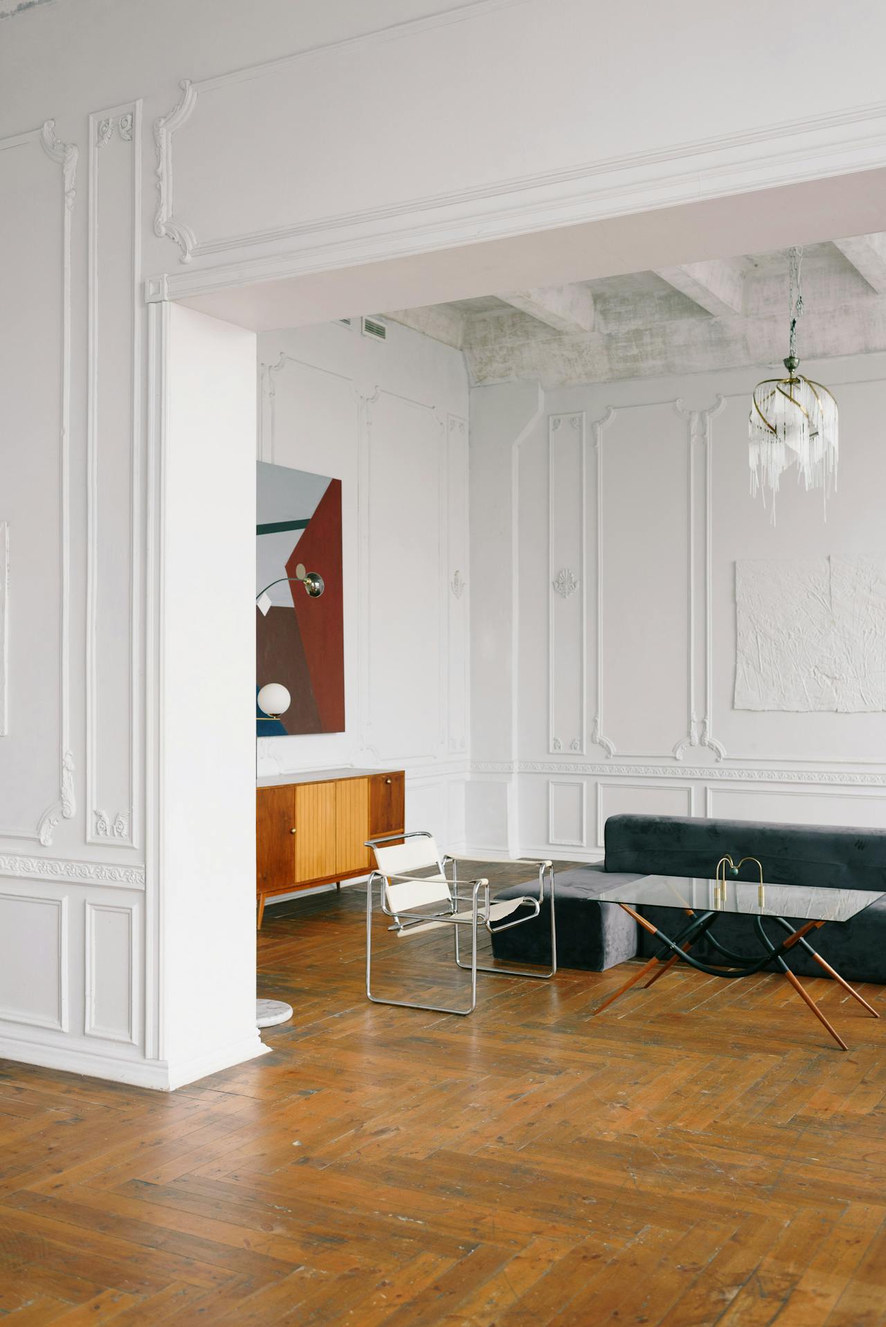 Minimalist living room with wood floor, white walls, modern chair, dark sofa, glass coffee table, and abstract artwork.
