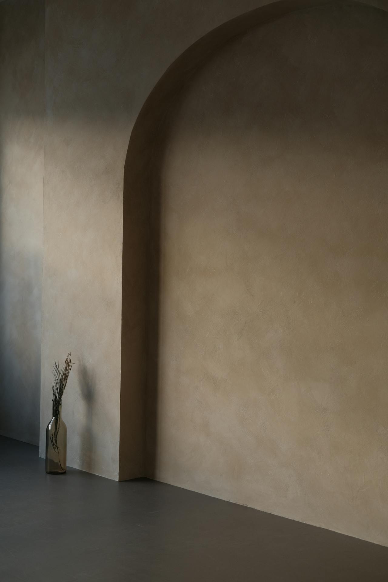 Minimalist interior wall with an arched niche and a glass vase holding dried plants on the floor.