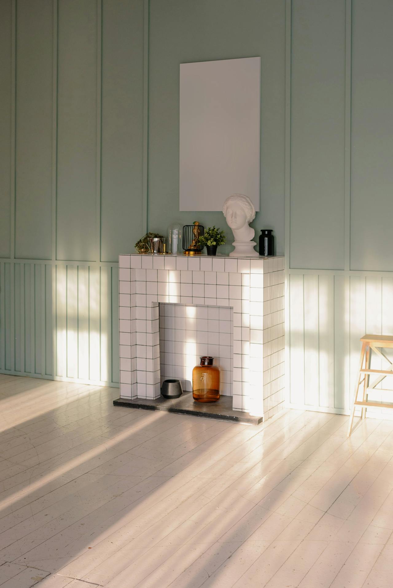 Sunlit white tiled fireplace with decorative bust, plants, vases, and blank wall art on green paneled wall.