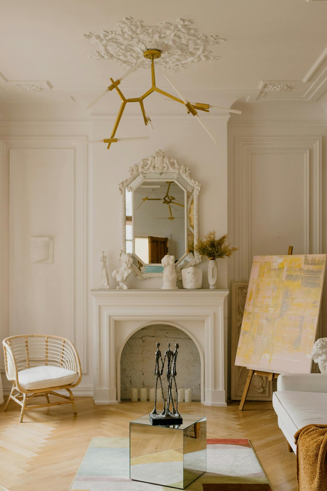 Minimalist living room with rattan chair, fireplace, mirror, sculpture, and abstract painting on easel.