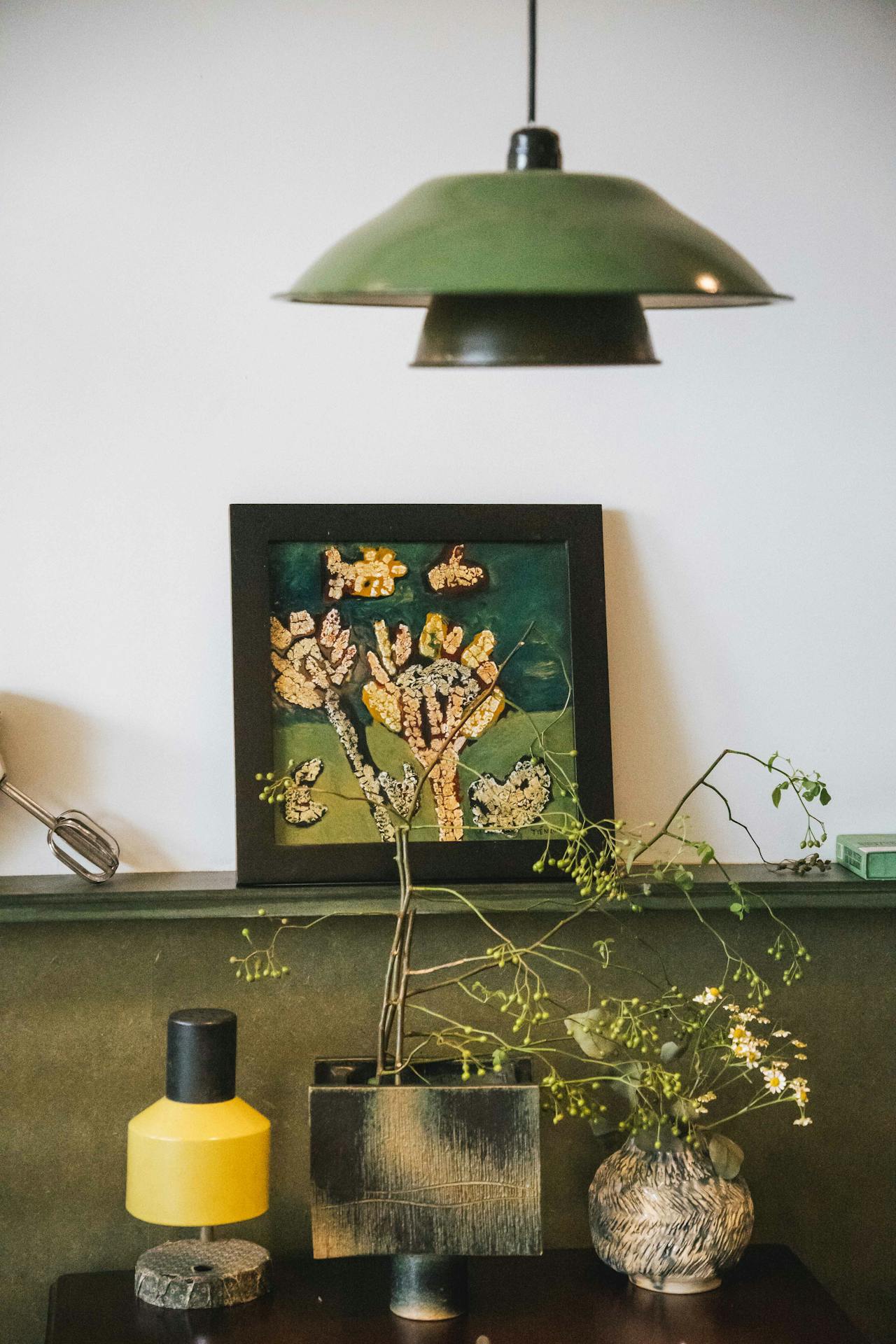 Interior with a green hanging lamp, artwork of abstract flowers, two vases with branches and flowers.
