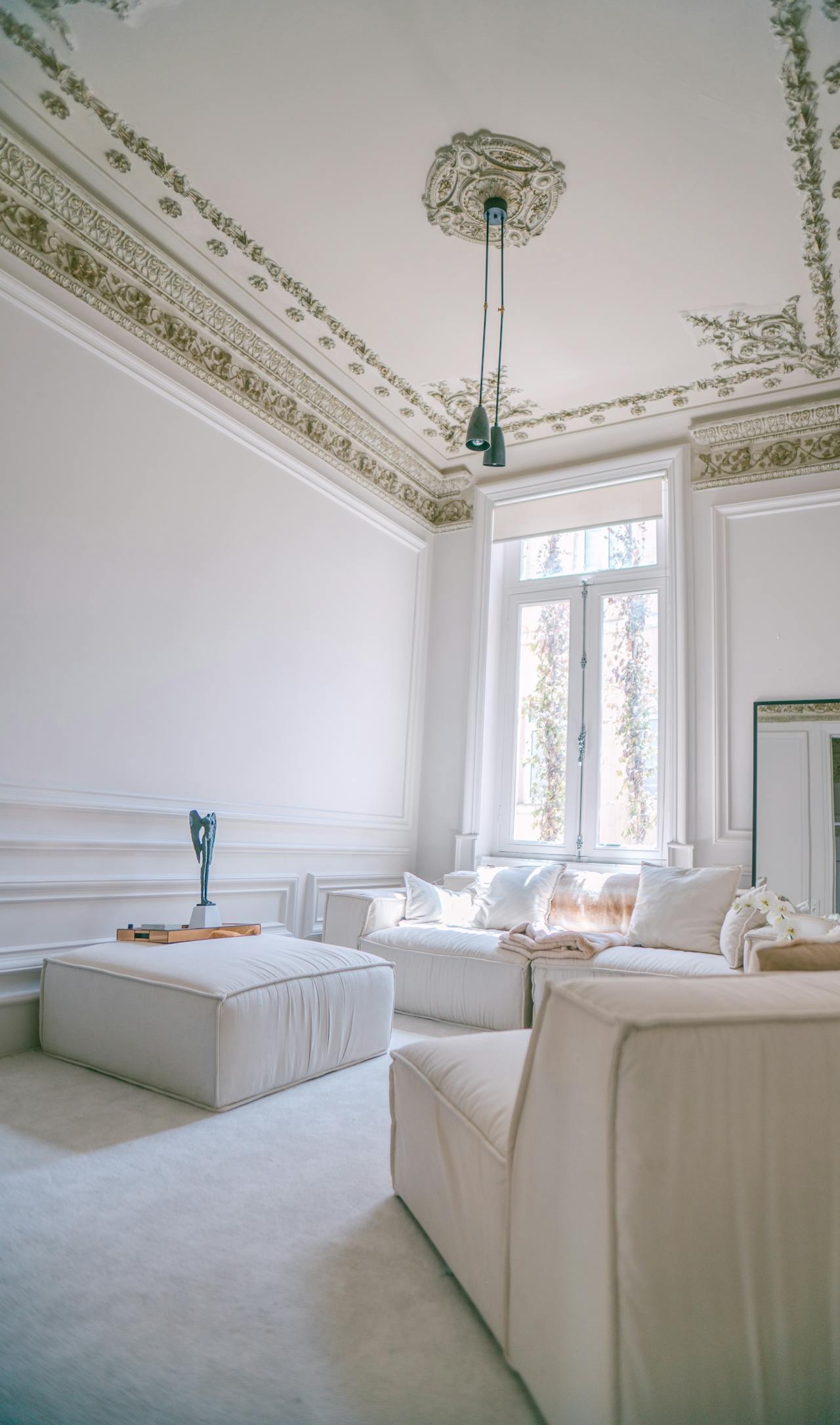 Bright living room with ornate ceiling, white sofa, armchair, ottoman, and large window with natural light.