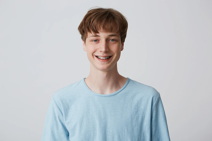 Teen boy with braces smiling and wearing a light blue t-shirt against a plain light gray background.