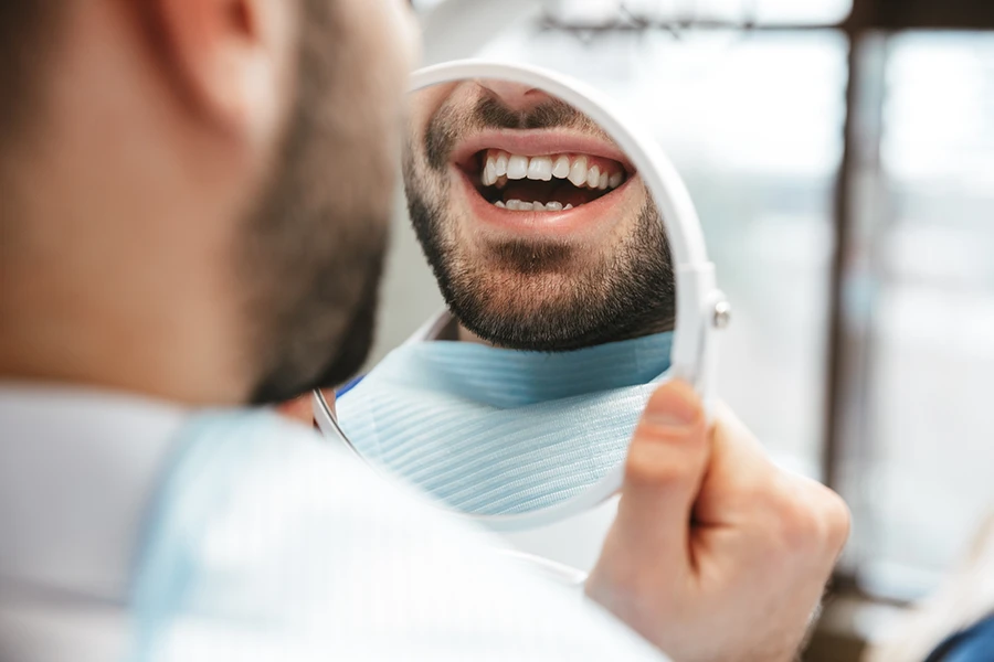 Bearded man smiling and looking at his teeth in a handheld round mirror wearing a dental bib.
